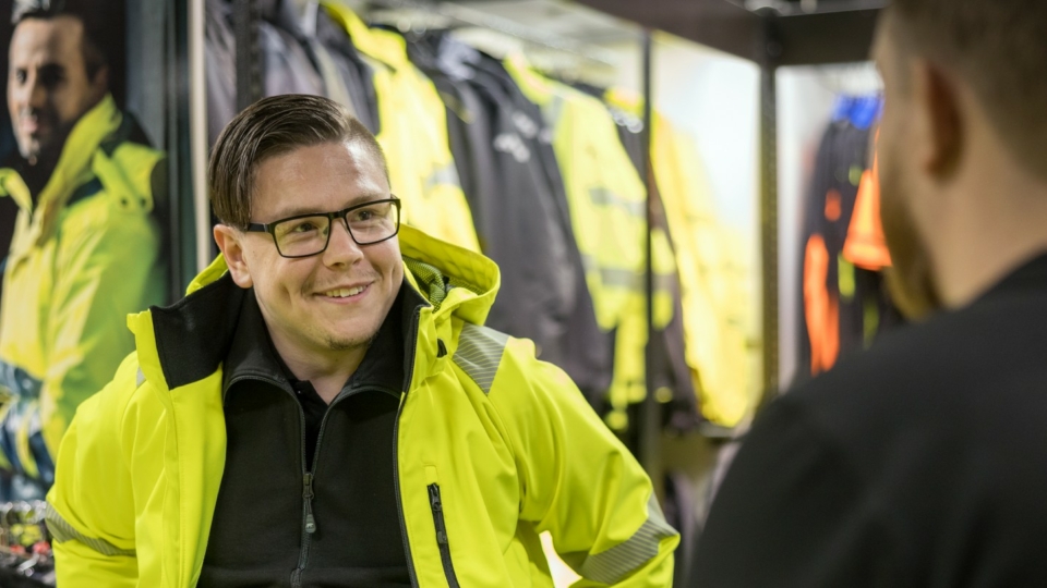 Two workers in yellow safety vests smiling in a warehouse with shelves in background
