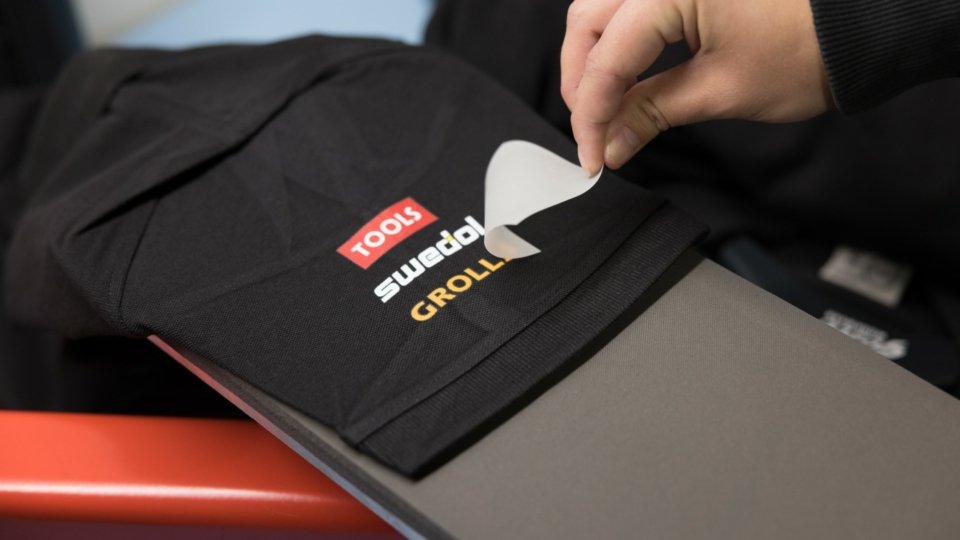 Hand peeling off transfer paper from black garment with TOOLS, Swedol, Grolls logos
