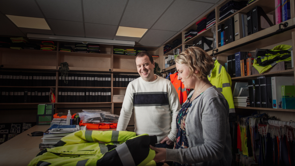 Two people examine high-visibility workwear in a showroom filled with samples and fabric binders