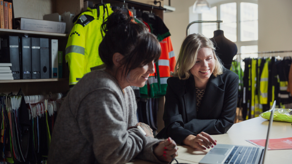 Two women smiling while looking at a laptop in a showroom with safety workwear