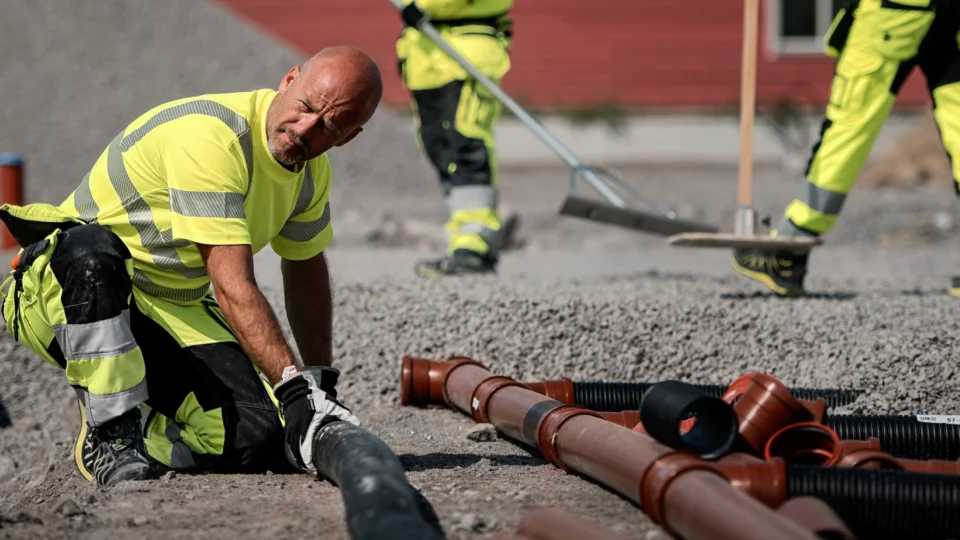 Construction worker installs black pipe in gravel while others rake the surface