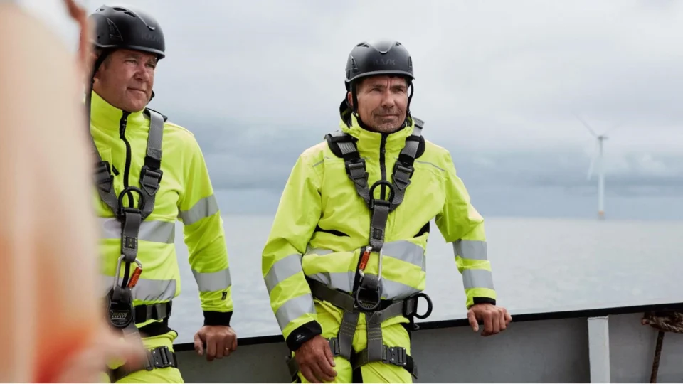Two workers in safety gear and helmets stand on a boat near an offshore wind turbine