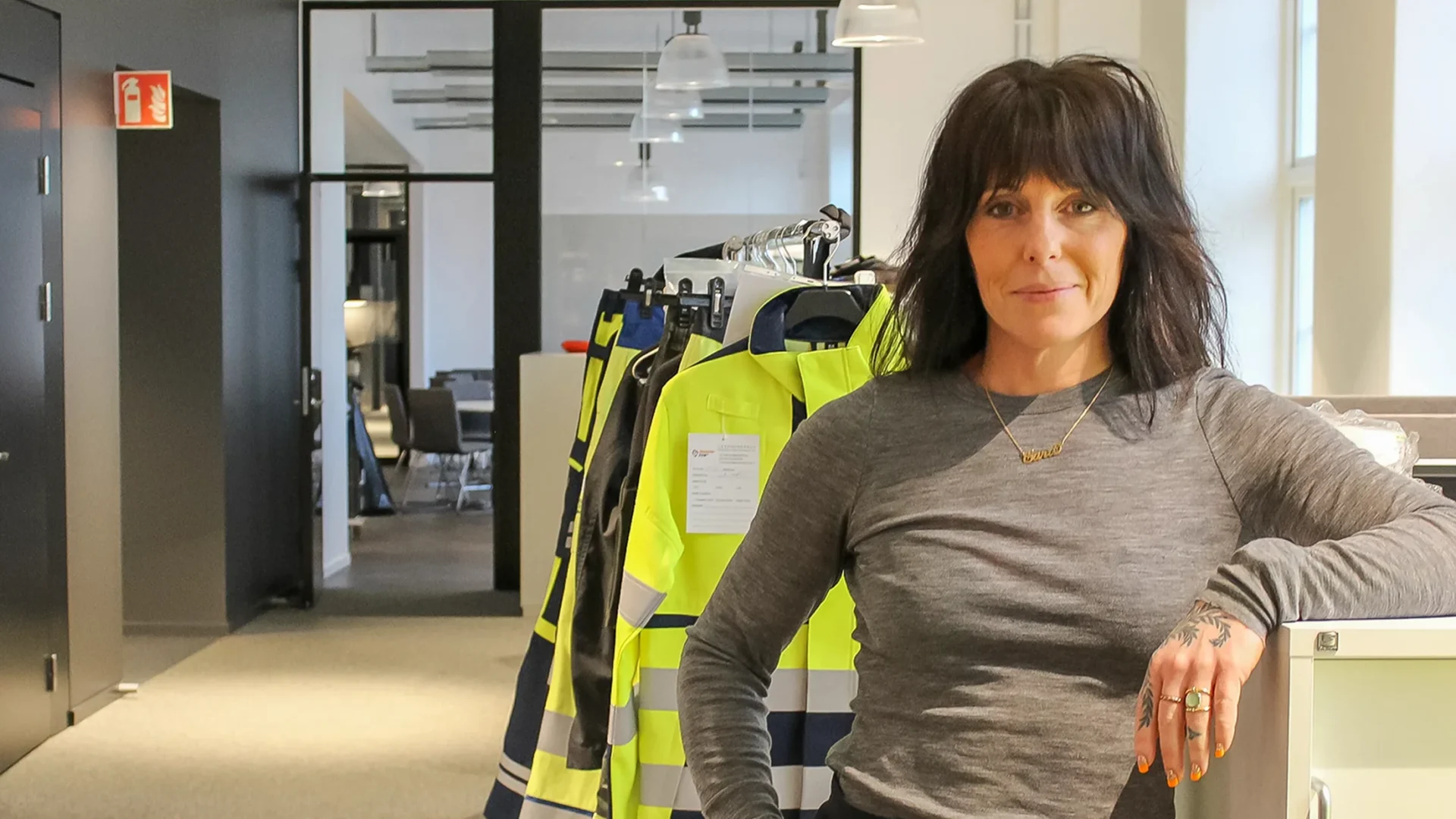 Sara Nygren Jönsson står på kontor bredvid upphängda varselkläder